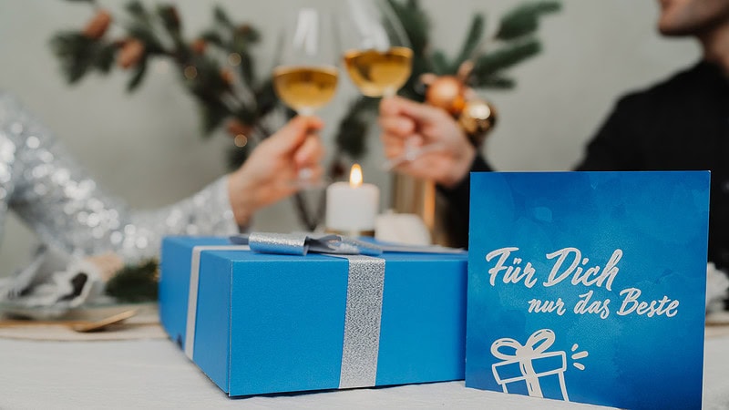 Ein Paar stößt mit einem Glaswein an. Im Vordergrund liegt eine Geschenkbox.