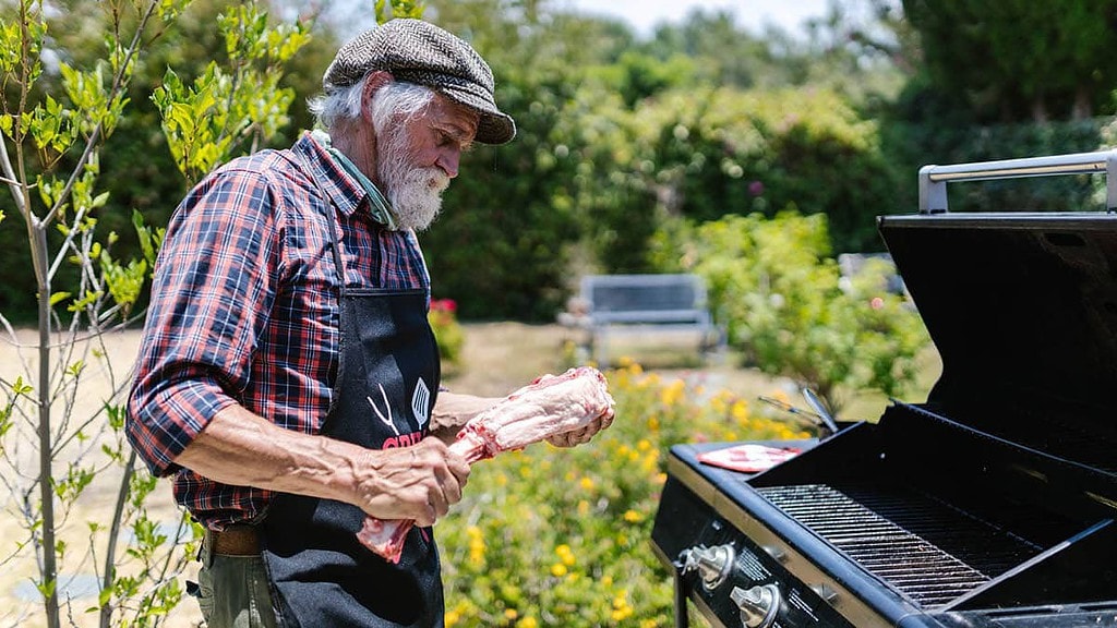 Grillviertell32.jpg Ein Mann steht neben einem Grill und hält Fleisch in der Hand