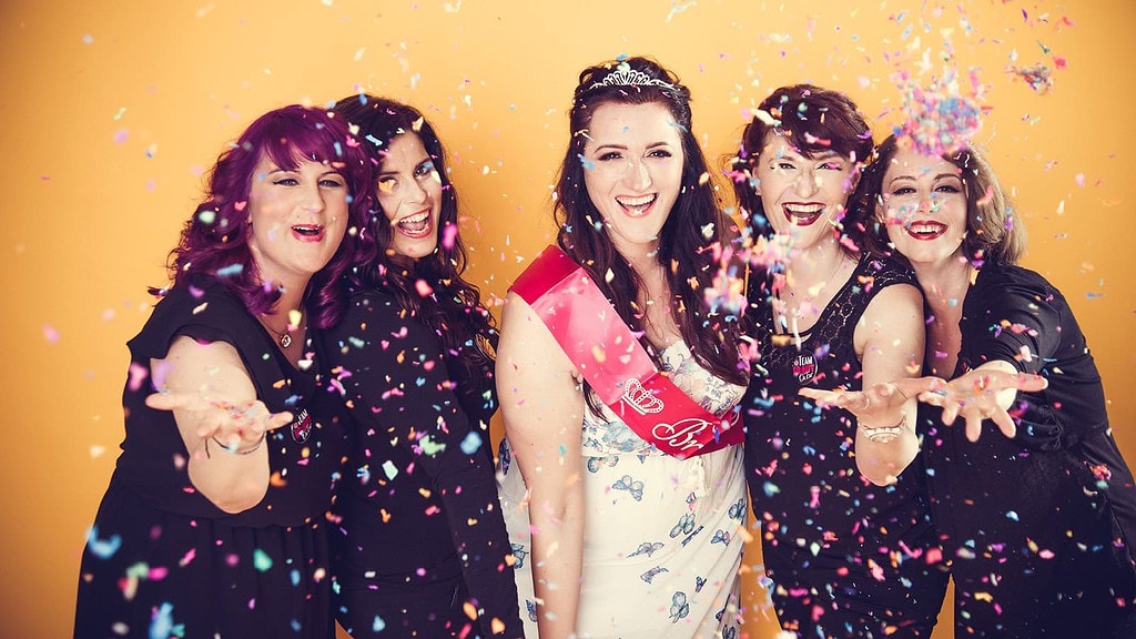 5 Frauen werfen Konfetti und lachen. eine hat ein weißes Kleid an