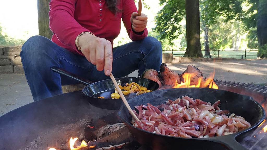 Teamevent Kochen am Lagerfeuer Frau am Lagerfeuer mit zwei Pfannen und Gemüse darüber Frau am Lagerfeuer mit zwei Pfannen und Gemüse darüber
