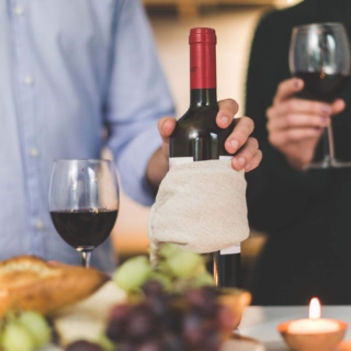 Ein Mann hält eine Rotwein-Flasche und ein Glas, eine Frau daneben trinkt den Rotwein