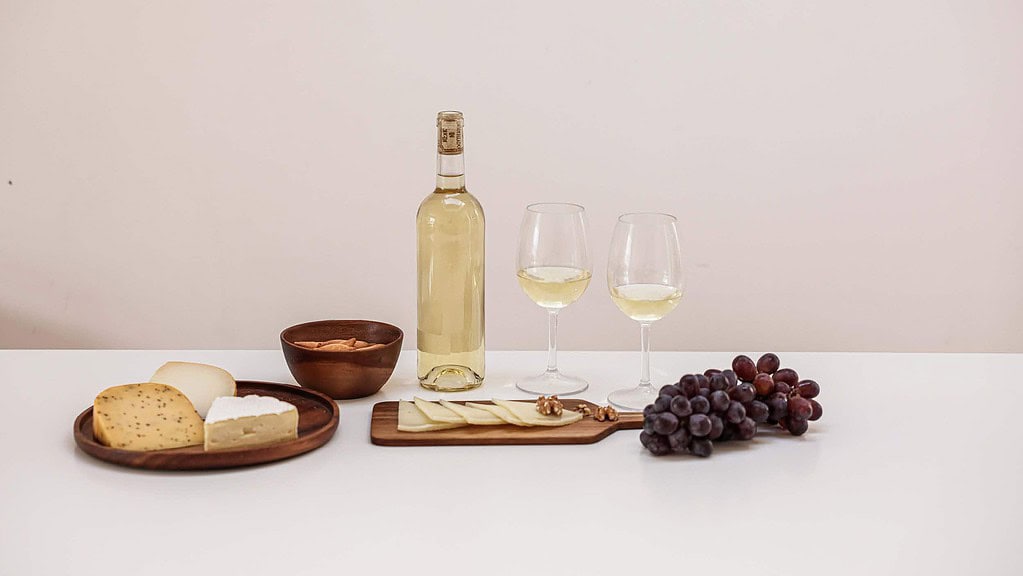Wein8.jpg Eine Weißweinflasche, zwei Weingläser stehen auf einem Tisch, daneben Käse, Brot und Trauben