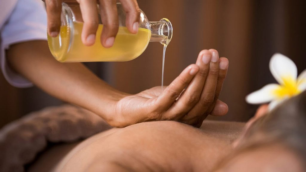 ayurvedische-massage Die Masseurin gibt eine ayurvedische Massage und hält eine Flasche mit Massageöl in der Hand.