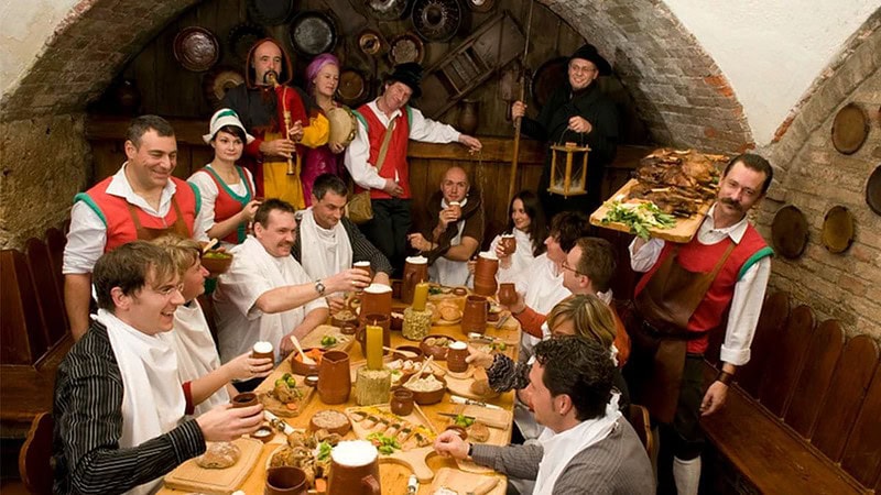 Die Teilnehmer des Ritteressens in München sitzen an der Tafel. Daneben Künstler und Kellner.