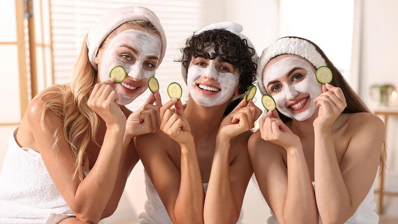 3 Freunde verbringen gemeinsam Zeit in einem Spa, tragen Handtücher, Gesichtsmasken und halten Gurkenscheiben in den Händen.