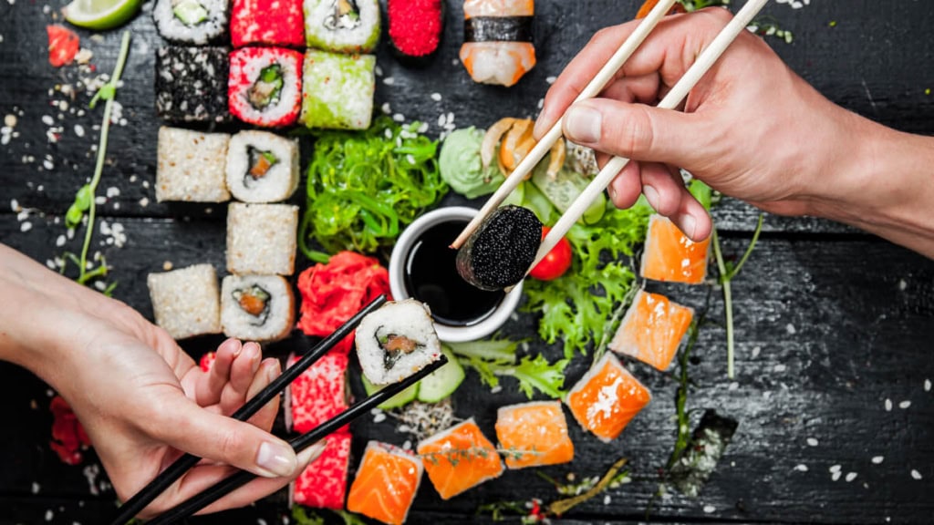 sushi-abend-dresden (2) Großer dunkler Teller mit Sushi-Stücken und Toppings wie Ingwer und Wasabi, 2 Hände über dem Teller, die das Sushi mit Stäbchen halten