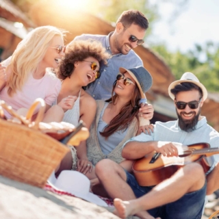 Eine Gruppe von Freunden macht ein Picknick, ein Mann spielt auf der Gitarre, die anderen lachen.