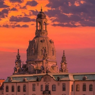 Die Skyline von Dresden wird bei einem rot-lilanen Sonnenuntergang dargestellt. Im Mittelpunkt steht vor allem die Frauenkirche.