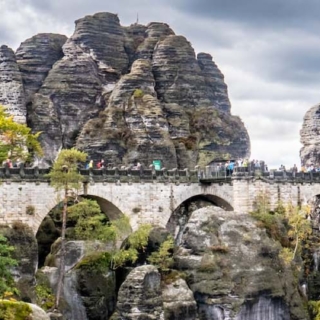 Im Mittelpunkt steht die Basteibrücke der sächsischen Schweiz. Man sieht viele grüne Bäume am Rand des Bildes.