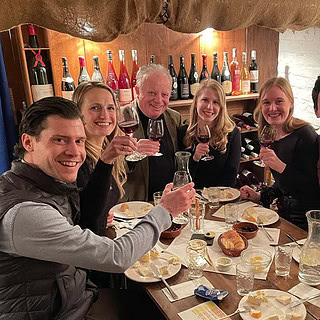 Eine Gruppe aus 7 Personen die mit einem Wein anstoßen, an einem gedeckten Tisch mit Käse Platten