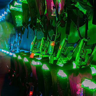 Lasertag Westen und Gewehre die an einer grün Leuchtenden Halterung an der Wand hängen.