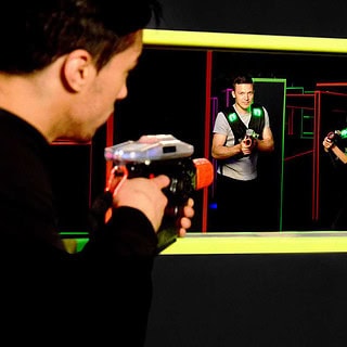 Die Sicht eines Lasertag Spielers der aus einem Fenster mit Gelben Ramen auf zwei andere Lasertag Spieler zielt die auch auf ihn zielen.