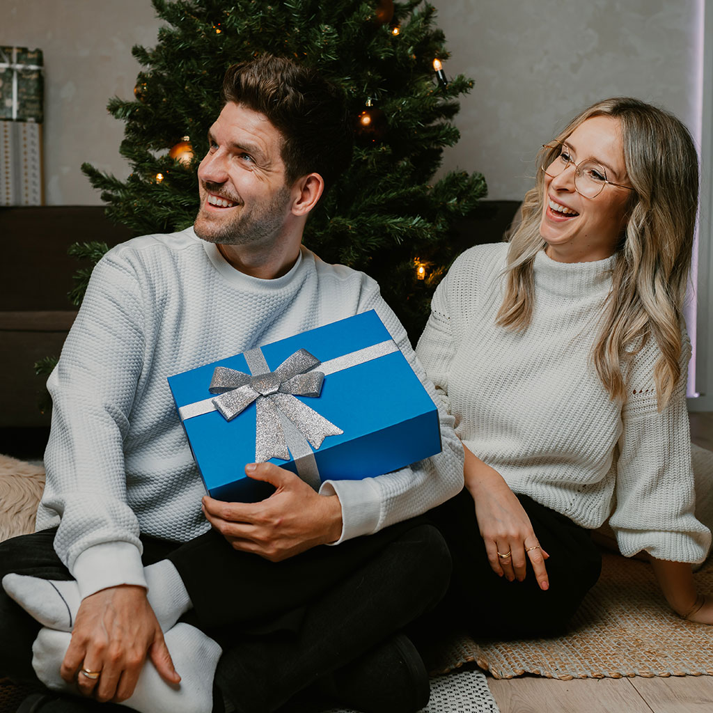 geschenkbox-blick-links Mann und Frau mit der Geschenkbox der Erlebnisfabrik in der Hand schauen nach links, im Hintergrund befindet sich ein Weihnachtsbaum