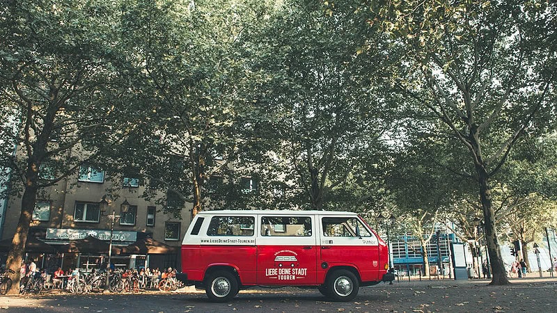 VW Bulli der vor Bäumen einer Allee parkt.