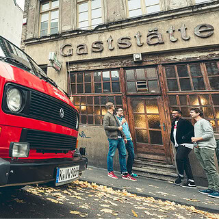 VW Bulli der vor einer Gaststätte parkt. Lächelnde Leute stehen vor dem Eingang der Gaststätte.