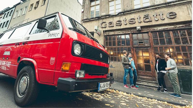 VW Bulli der vor einer Gaststätte parkt. Lächelnde Leute stehen vor dem Eingang der Gaststätte.