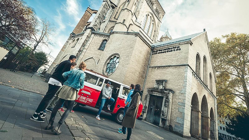 VW Bulli der vor einer Kirche parkt. Eine Gruppe steht vor dem Bulli.