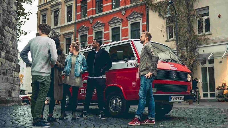 Eine Gruppe die neben einem VW Bulli steht und sich ein Gebäude anschaut.