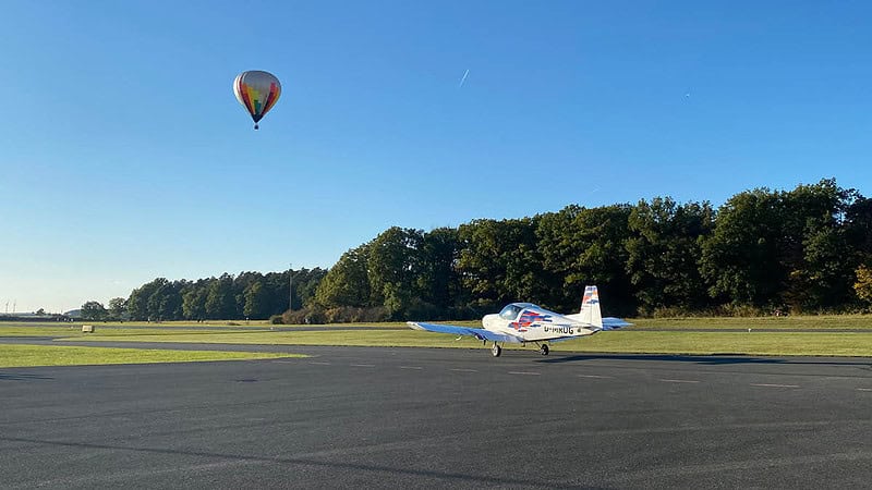 Ein Flugzeug steht bereit zum Abflug. Im Hintergrund fliegt ein Heissluftballon.