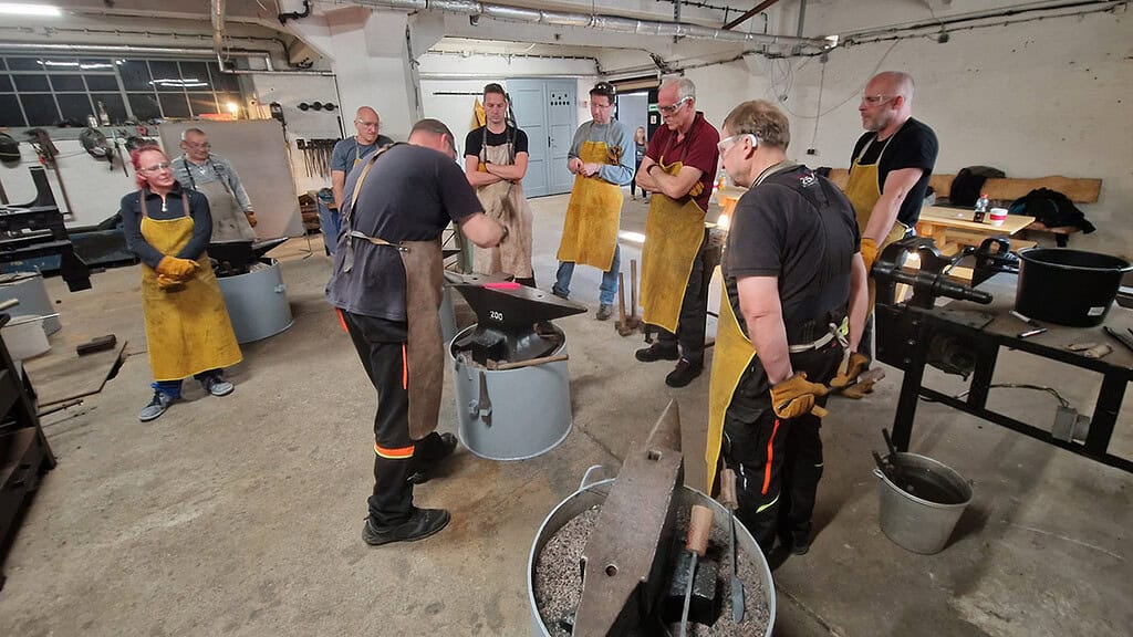 Der Kursleiter erklärt wie man genau schmiedet, damit alle es richtig machen. Person steht in der Mitte und schmiedet, alle andere haben die gelbe Schürze an und schauen aufmerksam zu.