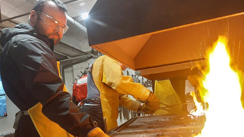 Eine Person mit Schutzbrille steht vor dem offenen Feuer an dem die Werkstücke hocherhitzt werden.