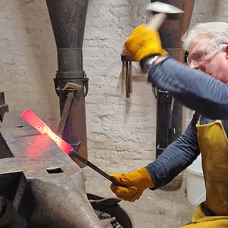 Ein Mann schlägt und Formt das Metall, um es später als Messer zu verarbeiten und zu veredeln. Das Bild ist ein einer Schmiede aufgenommen.