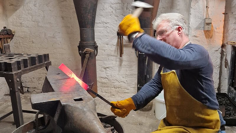 Ein Mann schlägt und Formt das Metall, um es später als Messer zu verarbeiten und zu veredeln. Das Bild ist ein einer Schmiede aufgenommen.