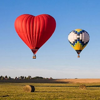 Roter herzförmiger Ballon neben zwei bunten Ballonen die über einem Feld fliegen- Ballonfahrt- Ballons über Feld