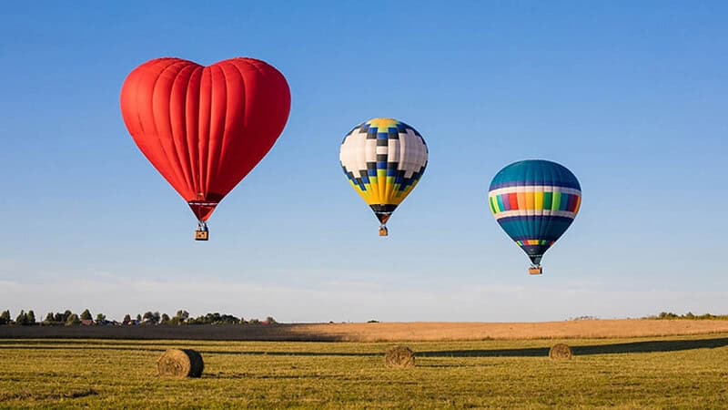 Roter herzförmiger Ballon neben zwei bunten Ballonen die über einem Feld fliegen- Ballonfahrt- Ballons über Feld