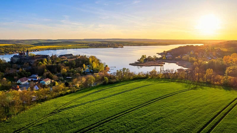Aussicht auf Landschaft mit Waldabschnitt und See, im Hintergrund Sonnenaufgang- Ballonfahrt- Landschaft mit Sonnenaufgang