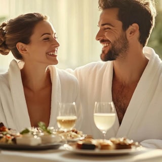 Ein lachendes Paar im Bademantel welches an einem gedecktem Tisch sitzt.